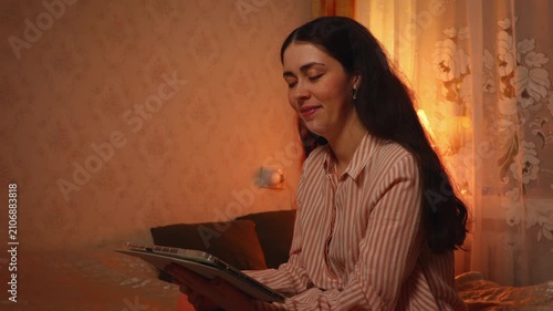 Side view of a young Caucasian woman relaxing and smiling while watching an online video on a tablet in the evening. Digital entertainment, home leisure, and modern lifestyle concept.