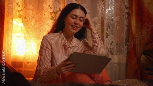 Beautiful young Caucasian woman with braces relaxing and smiling while watching an online stream on the internet in the evening. Digital entertainment, home leisure