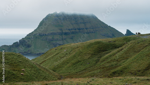 Grazing sheep on rolling green hills with distant hikers standing on a ridge, looking out toward the cloud-capped islet of Tindholmur across the ocean on Vagar Island, Faroe Islands