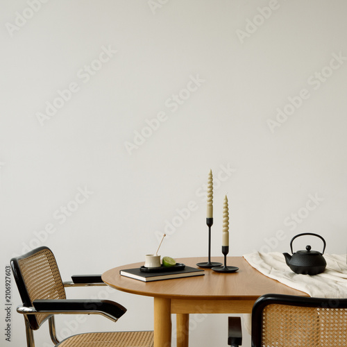 Minimalist dining table with yellow flowers in glass vase, candles, black teapot, and limes, set against a white wall. Copy space.
