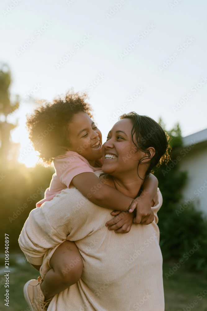 Fototapeta premium Laughing woman giving a piggyback ride to a small child in a sunlit backyard, featuring hazy summer light and soft ribbed textures for family bond and parenting lifestyle content