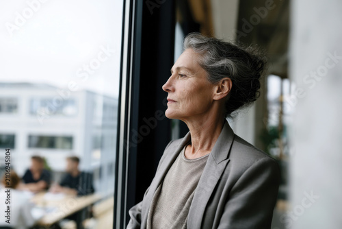 An older woman reflects deeply while gazing out the window in a modern office setting, showcasing elegance and introspection in her stylish gray attire.