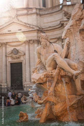 Sculpture of a river deity on Bernini's Fountain of Four Rivers, showing ancient art and architecture with a busy street scene in the background on a sunny day in the historic Italian capital