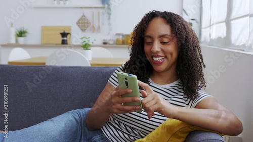 Young African American woman relaxing on sofa while using smartphone at home. Smiling young woman laughing while browsing social media content in a modern apartment. Technology and lifestyle concept.