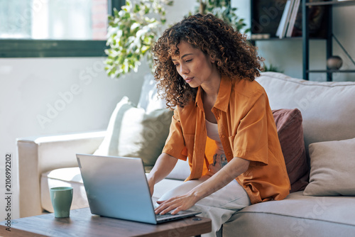 Obraz na plátně Concentrated woman working with her laptop while sitting on a couch at home