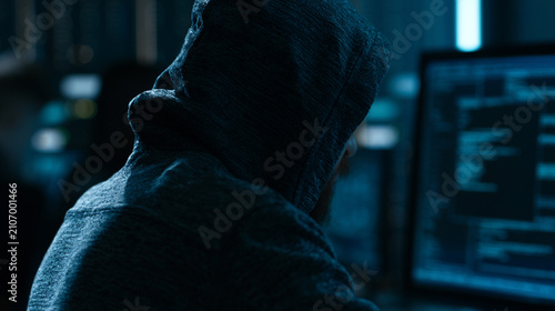 A mysterious figure in a hoodie sits in front of multiple computer screens, immersed in coding and cyber activities. The dark environment highlights a tech-driven world.