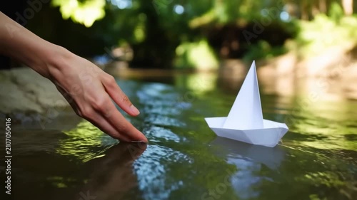 Hands of a person gently placing a white paper boat onto the surface of a serene stream surrounded by lush greenery and natural sunlight filtering through trees