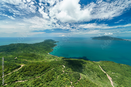 Lush green hills covered with tropical forests meet the vast blue ocean waters of the East Sea under a bright sky with white clouds during a sunny afternoon