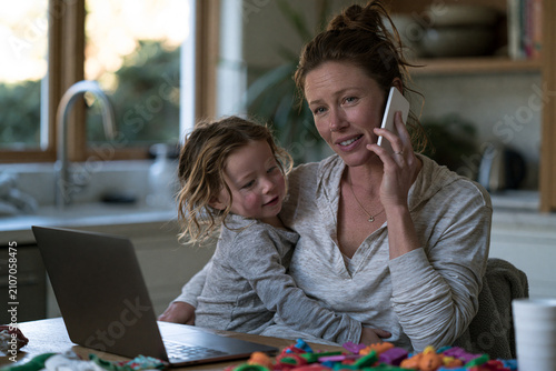 Woman with child working from home while talking on smartphone near laptop. Business mother multitasking with kid on lap in kitchen. Modern lifestyle of freelancer busy with job.