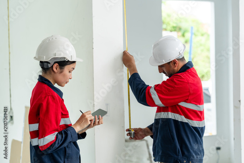 Asian female engineer checks tablet report while male contractor measures wall line with tape, indoor construction site safety inspection, quality control, building renovation progress review