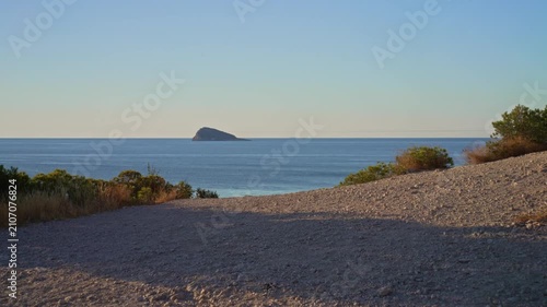 The camera shows the ocean and an island in the distance during sunset. The sun sets over the water while waves reach the shore with some plants around.