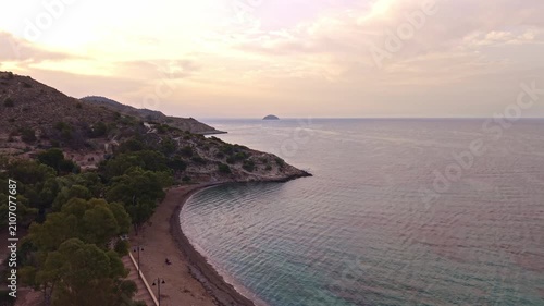 The video shows a coastal area with a beach and water. The sun sets over the mountains and the beach. Soft colors fill the sky. People walk along the shore.