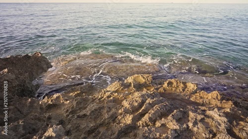 Waves come in and go out against the rocks by the shore during sunset. The sky is clear and the water moves lightly. This captures the beauty of nature at dusk.