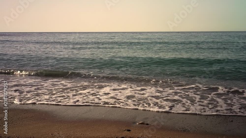 Waves crash softly on the sand at the beach. The sun sets over the horizon, creating a calm atmosphere. The sound of waves fills the air as the sky changes colors.