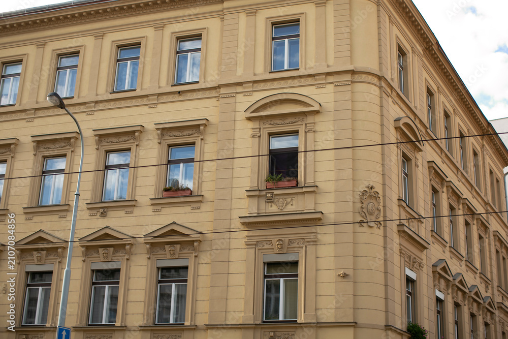 Fototapeta premium Ornate corner facade with window accents and urban details, soft golden plaster tones, decorative frames and subtle shadow interplay, small planter