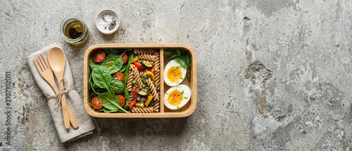 Obraz  z motywem Balanced lunchbox containing pasta salad and boiled eggs on stone surface. Top view of healthy meal prep with wooden cutlery and oil.