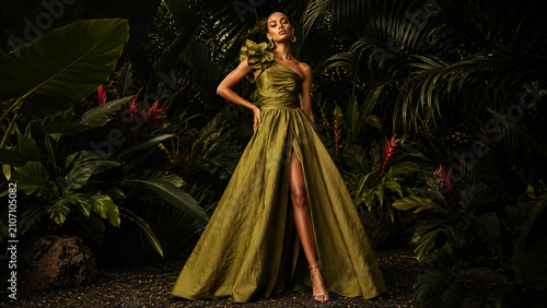 Elegant woman in a flowing olive gown posing in a dark tropical garden.