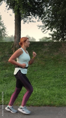 Vertical video. Young athletic woman with red hair running along a paved path in a city park during her morning workout, maintaining a healthy and active lifestyle with cardio exercises and training