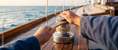 Sailor hands securing rope on winch aboard wooden deck of yacht at sea during sunset