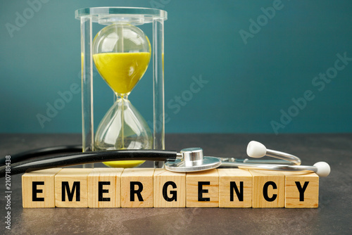 Obraz na plátně Hourglass and stethoscope above wooden blocks spelling EMERGENCY on a dark surface