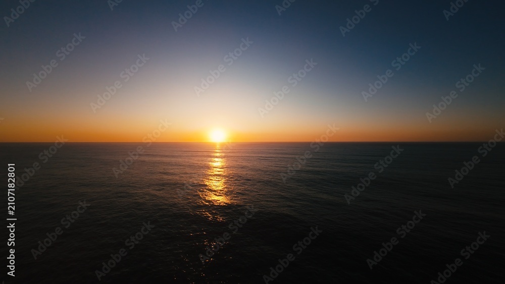 Obraz premium Aerial view of sun dipping into ocean, calm waters shimmering with golden light
