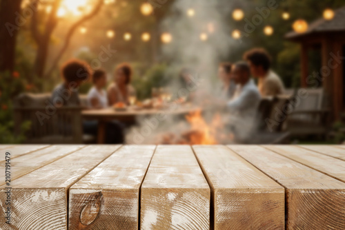 Empty wooden table for product placement. Blurred background with friends and family gathered in garden enjoying summer barbecue on sunny day. 