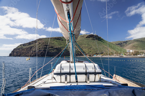 Schilderij op canvas The bow of a sailing yacht approaching the green headland and black sand bay of