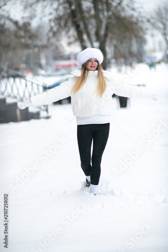 Happy young woman posing outdoors in winter snow, wearing a cozy white puffer jacket, white mittens, and a black knit beanie. Winter fashion concept.