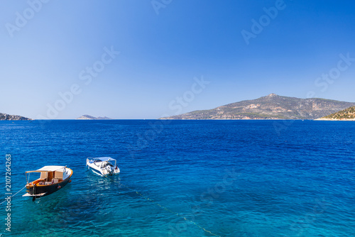 Canvas-taulu Two small boats tethered on a vibrant blue sea with clear skies and distant island hills, evoking summer travel and peaceful coastal scenery ideal for vacation, leisure and nautical themes