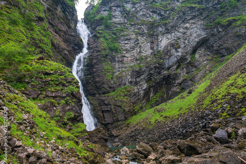Tall narrow waterfall flowing through a rocky green ravine in the Geiranger region, Norway.