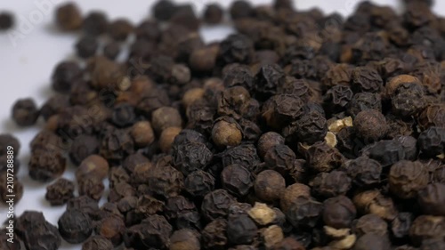 Close-up of a heap of dry black peppercorns rotating on a white plate, showcasing their wrinkled texture and natural brown tones under studio light.