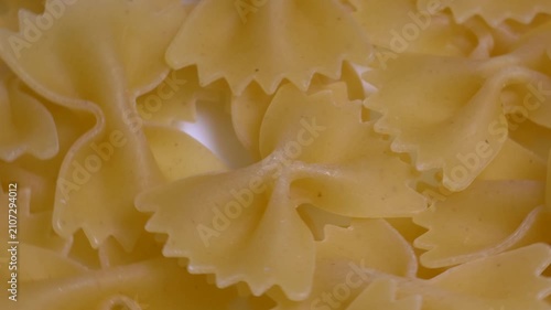 Close-up of raw farfalle bow-tie pasta rotating on a white plate, showcasing its textured shape and golden yellow color under bright studio lighting.
