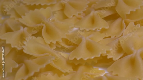 Close-up of raw farfalle bow-tie pasta rotating on a white plate, showcasing its textured shape and golden yellow color under bright studio lighting.