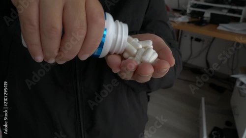 Close-up of a hand pouring white medical capsules from a bottle into an open palm, highlighting the daily health routine under soft lighting.