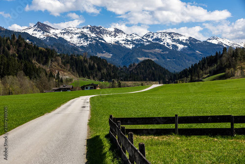 A path leading into the distance and a wooden fence in the spring Tyrolean Windautal valley, in the background snowy Alpine peaks under a blue sky with white clouds