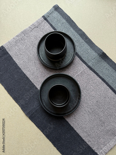 Two black ceramic cups on a diagonally placed linen napkin. Handmade stoneware with natural textile texture creates a dynamic minimalist composition for home decor, dining and interior concepts.