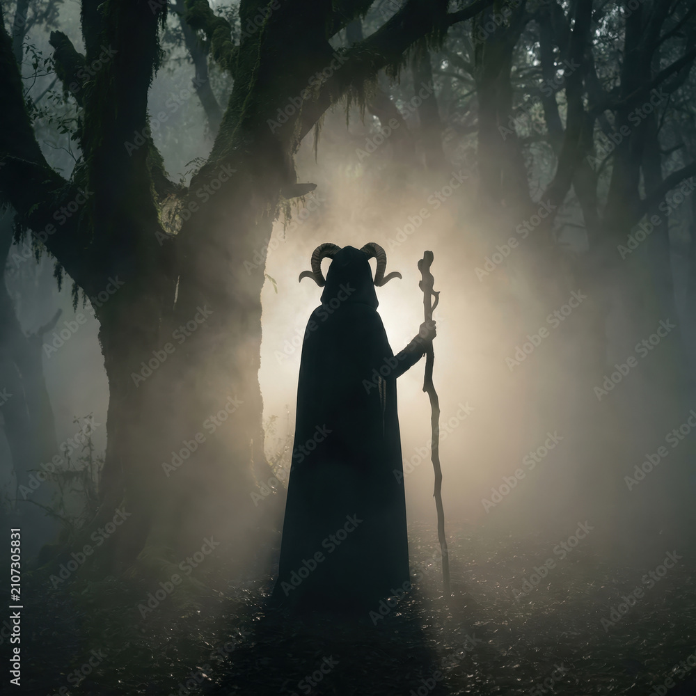 Obraz premium mysterious silhouetted figure in cloak and horned headdress holding wooden staff in foggy forest. pagan mythology, occult magic and dark fantasy concept. halloween background with copyspace.