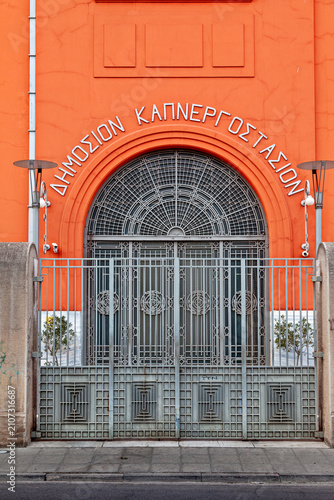 Ornate metal entrance of the historic Public Tobacco Factory in Athens, Greece, featuring Greek lettering, geometric ironwork and a vivid orange industrial façade. Nowadays it hosts part of the Parlia