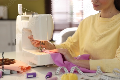 Woman sewing fabric with professional machine at white table in workshop, closeup