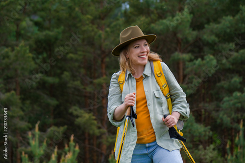Fotografie Smiling woman with trekking poles hiking in pine forest with yellow backpack