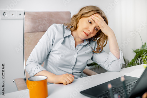 Stressed woman holding her head in pain at laptop desk in office. Workplace stress, headache concept, burnout syndrome.