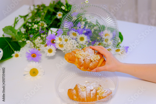 A fresh buttery croissant topped with powdered sugar is lifted from a clear takeaway container by a female hand. Crisp flaky layers create an inviting bakery breakfast ready to enjoy.