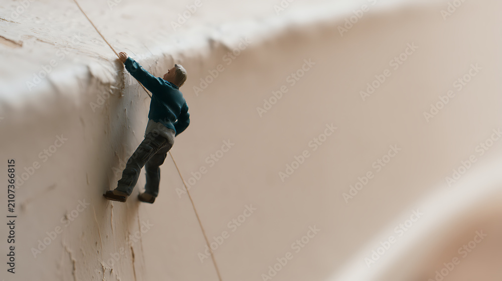 Fototapeta premium A small miniature figure, a person, climb a textured white and beige surface with a rope, illustrating challenge, perseverance, and effort