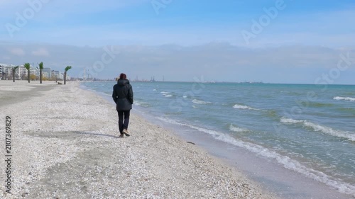 Lonely woman walking on a desolate winter beach. Solitary woman in a dark winter coat is walking away along a deserted seashell beach next to a cold sea, with a distant industrial port city visible