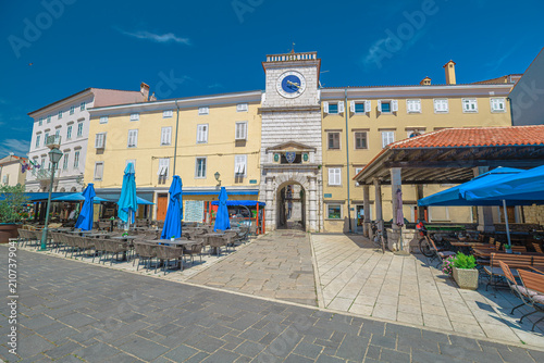 Cres town square, Trg Frane Petrica, presenting classic Venetian architecture and a prominent clock tower under a clear summer sky, with outdoor cafes inviting relaxation of Cres Island in Croatia.