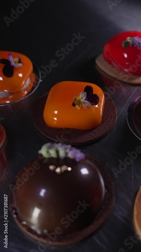 Close up of beautifully crafted assorted mousse pastries in chocolate, red, and orange with shiny mirror glazes and elegant decorations displayed on a table, haute cuisine confectionery