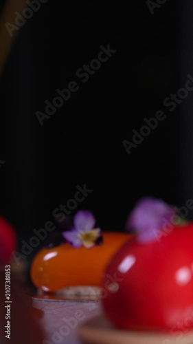Static shot with a rack focus pulling from a red to an orange mousse pastry, both featuring shiny mirror glazes and edible flower decorations, haute cuisine confectionery, cinematic food videography
