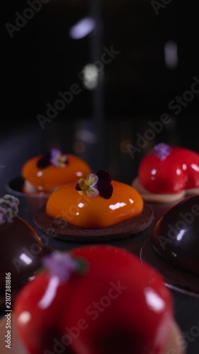 Close up of beautifully crafted assorted mousse pastries in chocolate, red, and orange with shiny mirror glazes and elegant decorations displayed on a table, haute cuisine confectionery