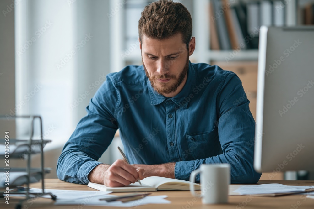 Obraz premium Man writing in a notebook at an office desk as a focused young male adult person