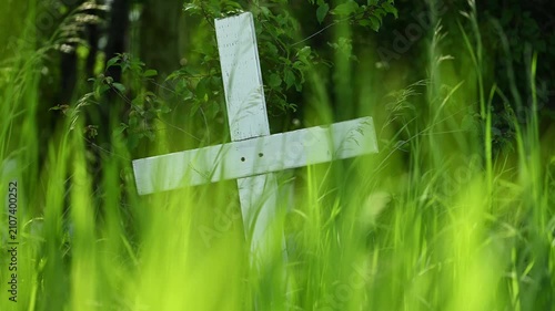 A weathered white wooden cross stands slightly tilted in a dense field of vibrant tall moving green grass and foliage.